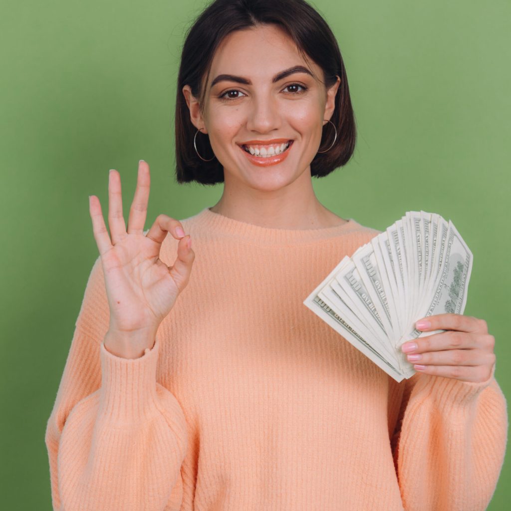 Young woman in casual peach sweater  isolated on green olive background  lucky holding fan of 100 dollar bills being excited to win cash prize showing OK sign